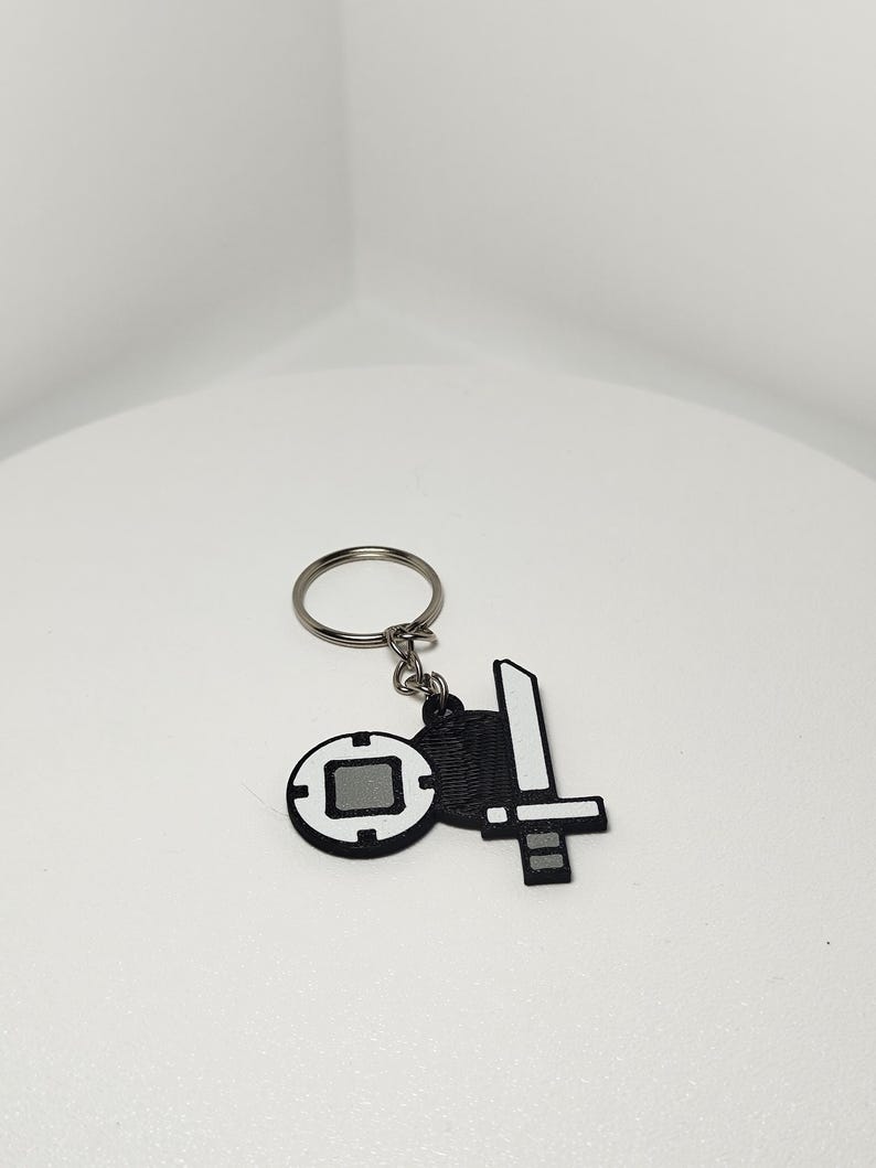 May include: A black and white keychain featuring two charms: a sword and a circular emblem. The sword charm has a white blade and a black handle. The circular emblem has a gray square in the center and a black border.