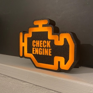 May include: A black and orange 3D printed sign shaped like a car's check engine light. The sign reads "CHECK ENGINE" in white letters.