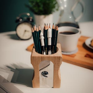 Op de afbeelding: Houten puntenslijper met potloden erin. De puntenslijper heeft een zilveren mes en een rechthoekige vorm. De potloden zijn groen en zwart. Een witte koffiemok en een kleine plant staan op de achtergrond.