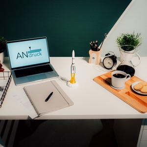 May include: A well-lit workspace featuring a laptop displaying the text "ANdruck", a rocket model, a notebook with a pen, and a cup of coffee with cookies on a wooden tray. Other items include a desk organizer, a small clock, and a wire basket.