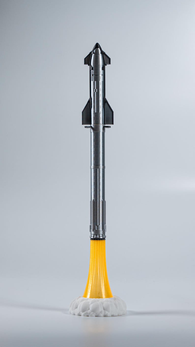 May include: A silver and black rocket model with a yellow flame effect at the base, resting on a white cloud-like structure. The rocket has a detailed design, with fins and a pointed nose. The model is a space-themed item.