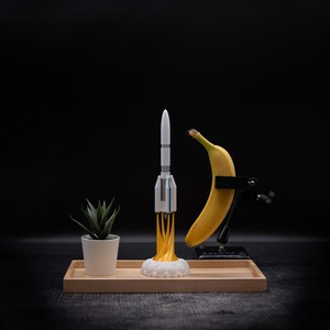 May include: A whimsical still life featuring a toy rocket with yellow flames, a yellow banana held by a clamp, and a small potted succulent. The items are arranged on a wooden tray against a dark background.