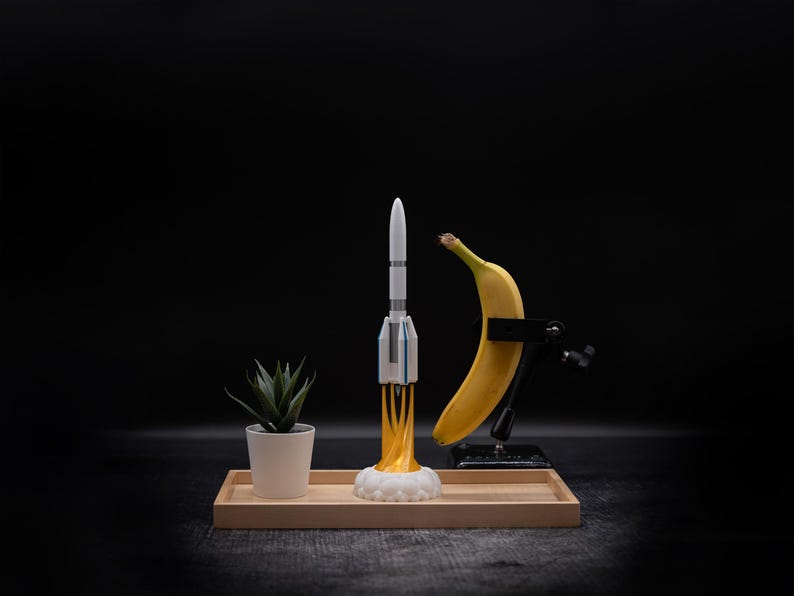 May include: A whimsical still life featuring a toy rocket with yellow flames, a yellow banana held by a clamp, and a small potted succulent. The items are arranged on a wooden tray against a dark background.