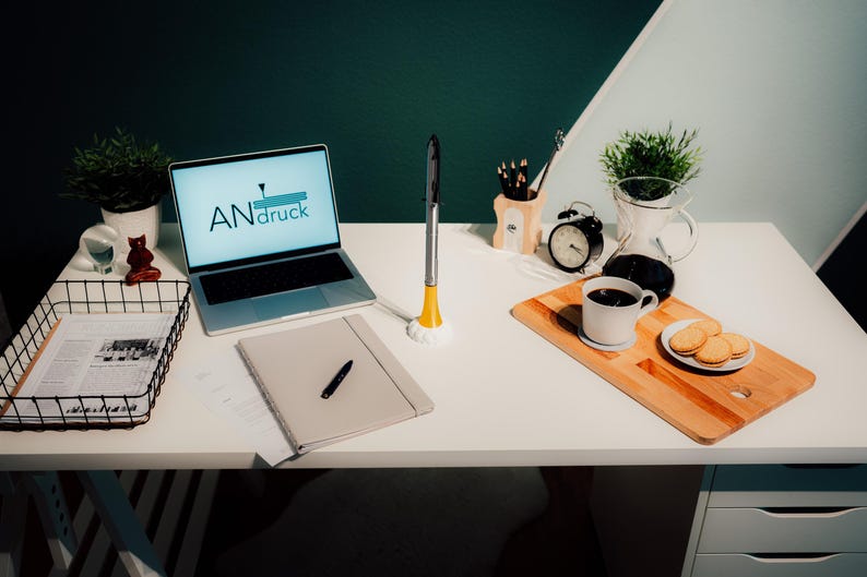 May include: A well-lit workspace featuring a laptop displaying "ANdruck" text, a notebook with a pen, and a coffee setup. A small plant, a clock, and a tray with cookies are also present. A unique desk accessory is also visible.