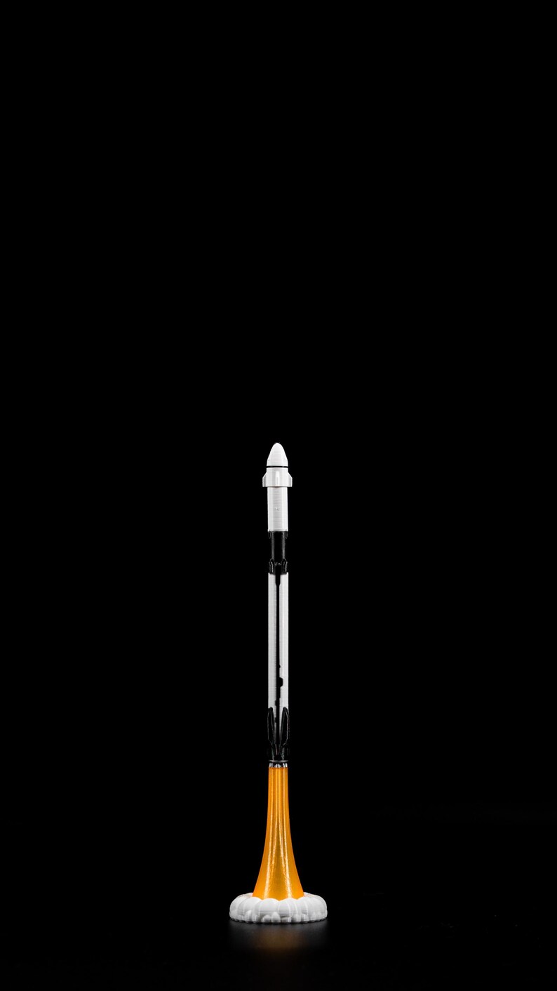 May include: A miniature rocket model against a black background. The rocket has a white nose cone, a black and white body, and an orange and white base, suggesting flames. The design is detailed and stylized.