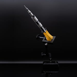May include: A model rocket, painted in black and white, is mounted on a black stand. The rocket has a pointed nose and is depicted with yellow flames emanating from its base, suggesting liftoff. The background is a dark, reflective surface.