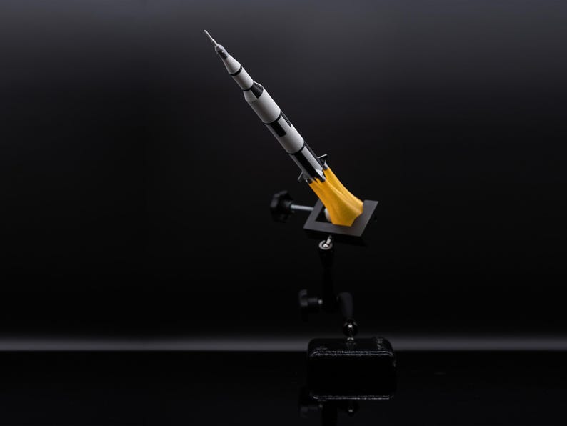 May include: A model rocket, painted in black and white, is mounted on a black stand. The rocket has a pointed nose and is depicted with yellow flames emanating from its base, suggesting liftoff. The background is a dark, reflective surface.