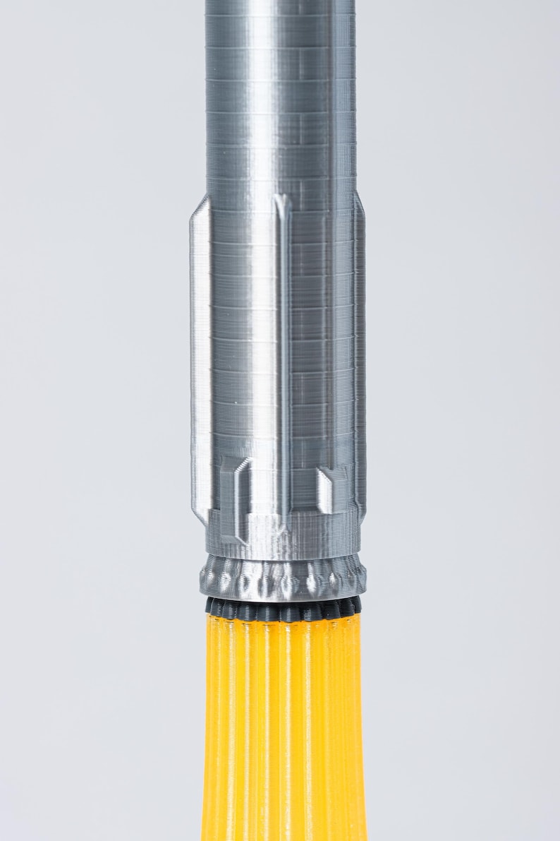 May include: A 3D-printed rocket model with a silver-gray upper section and a bright yellow lower section. The upper section has a textured surface with detailed paneling, while the lower section features vertical ridges. The rocket is set against a plain white background.