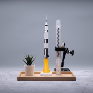 May include: A model rocket with black and white stripes and yellow flames is displayed on a wooden tray. A small potted plant and a ruler with a penguin figurine are also on the tray. The background is a solid gray.