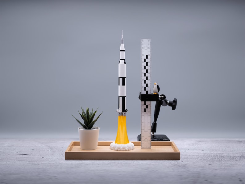 May include: A model rocket with black and white stripes and yellow flames is displayed on a wooden tray. A small potted plant and a ruler with a penguin figurine are also on the tray. The background is a solid gray.