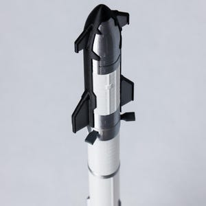 May include: A 3D-printed model rocket in black, white, and silver. The rocket has a pointed nose cone, fins, and a detailed engine section. The model is standing upright against a plain background.