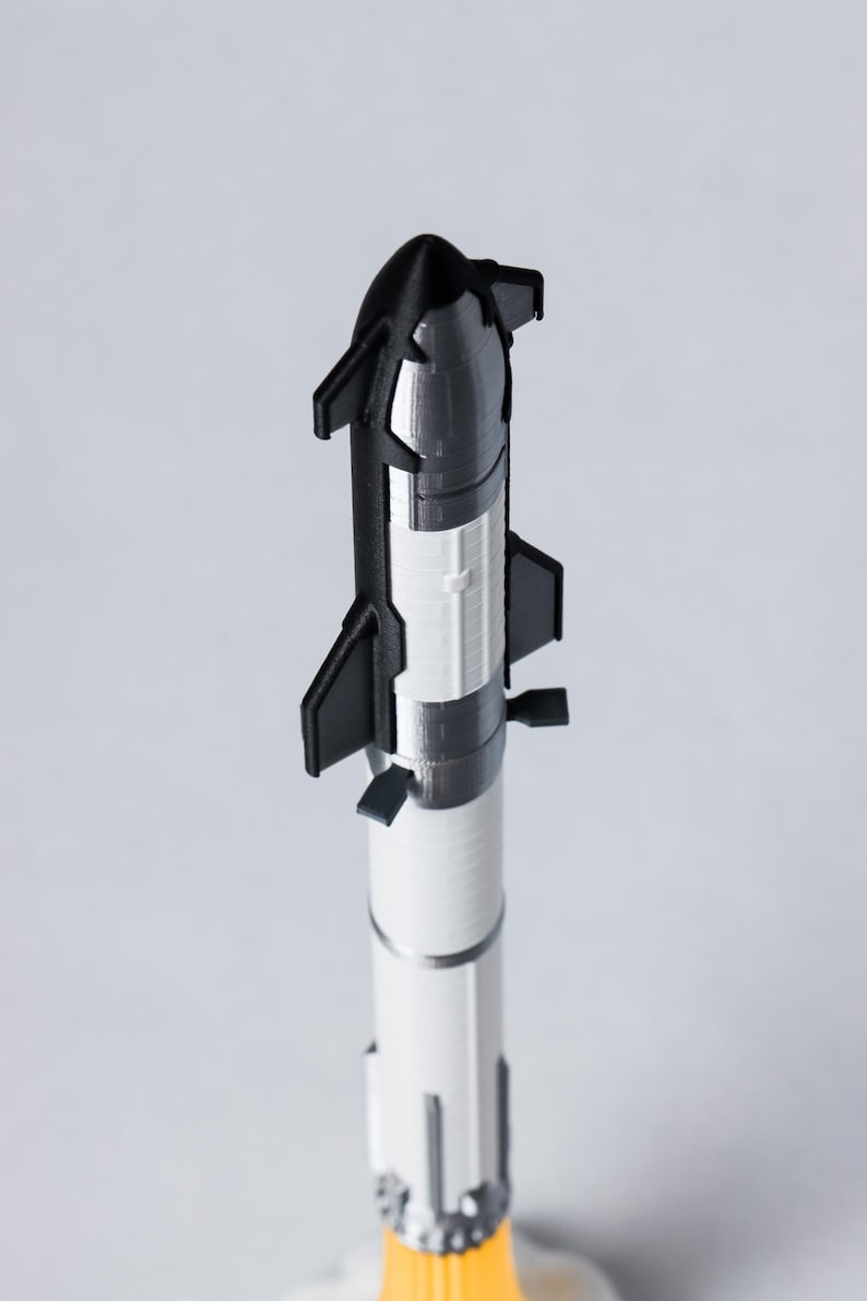 May include: A 3D-printed model rocket in black, white, and silver. The rocket has a pointed nose cone, fins, and a detailed engine section. The model is standing upright against a plain background.