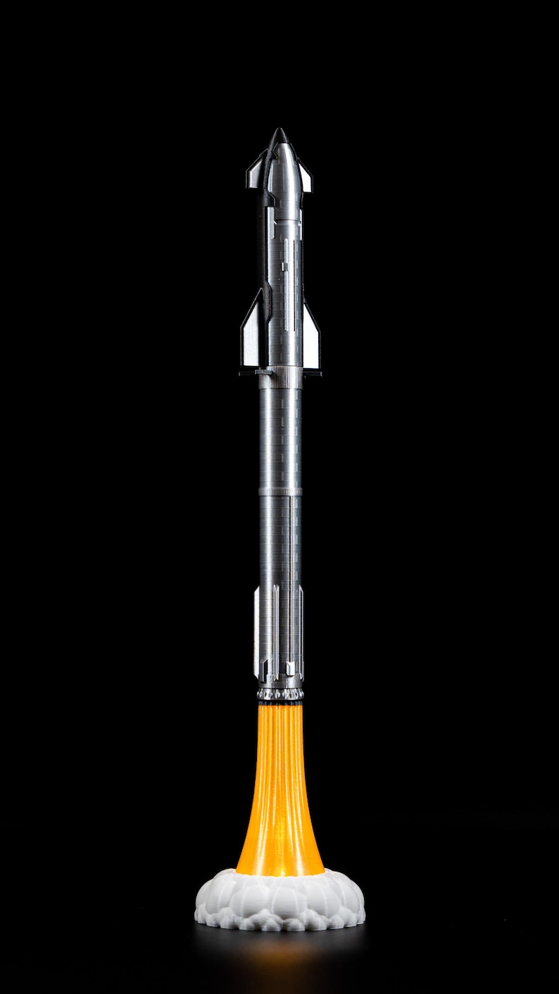May include: A silver rocket model with a pointed nose and detailed body, emitting a bright orange flame from its base. The rocket is set against a black background, with a white cloud-like base.