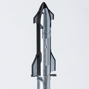 May include: A detailed model of a rocket, primarily silver with black accents. The rocket has a conical nose, fins, and a cylindrical body with ribbed detailing. The model is set against a plain white background.