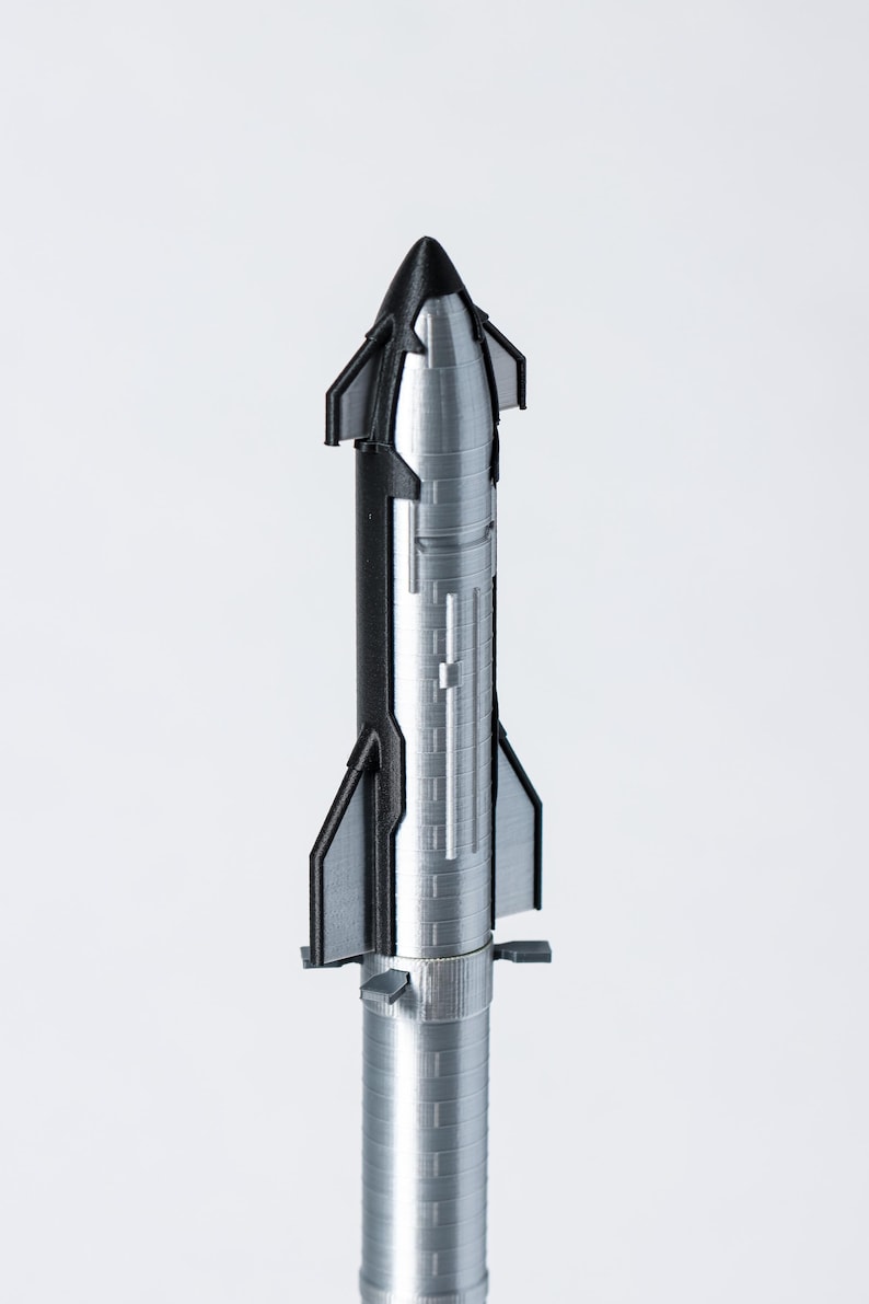 May include: A detailed model of a rocket, primarily silver with black accents. The rocket has a conical nose, fins, and a cylindrical body with ribbed detailing. The model is set against a plain white background.