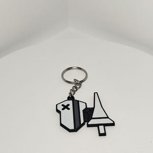 May include: A white and black keychain with a geometric design. The keychain features a silver key ring and chain. The design includes a white shape with a black X and a gray stripe, and a white shape with a black outline.