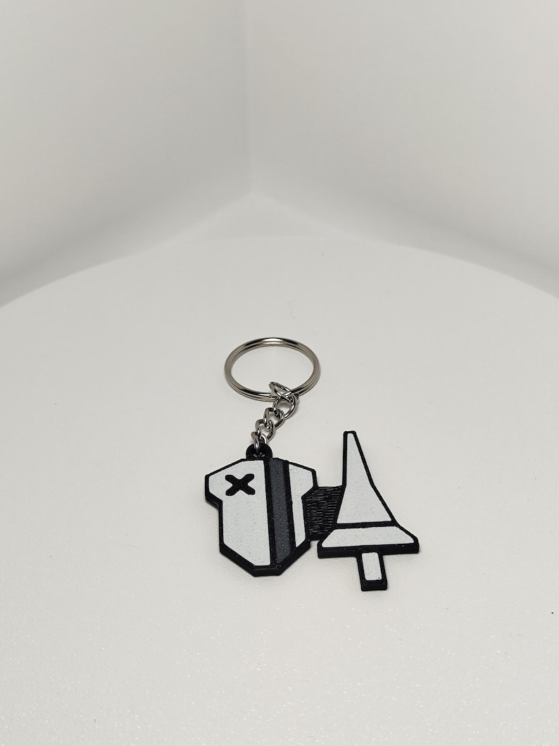 May include: A white and black keychain with a geometric design. The keychain features a silver key ring and chain. The design includes a white shape with a black X and a gray stripe, and a white shape with a black outline.