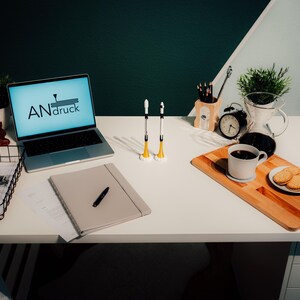 Op de afbeelding: Een goed verlicht bureau met een laptop met de tekst "ANdruck" op het scherm, een notitieboek, pennen en een koffiepauze met koekjes. Twee raketvormige objecten staan rechtop. Een draadmand bevat papieren.