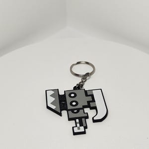 May include: A pixelated, gray and white keychain featuring a stylized axe design. The keychain has a silver metal ring for attaching to keys or bags. The axe design has a geometric, blocky appearance.
