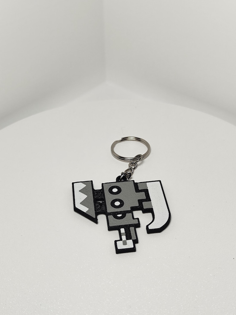 May include: A pixelated, gray and white keychain featuring a stylized axe design. The keychain has a silver metal ring for attaching to keys or bags. The axe design has a geometric, blocky appearance.