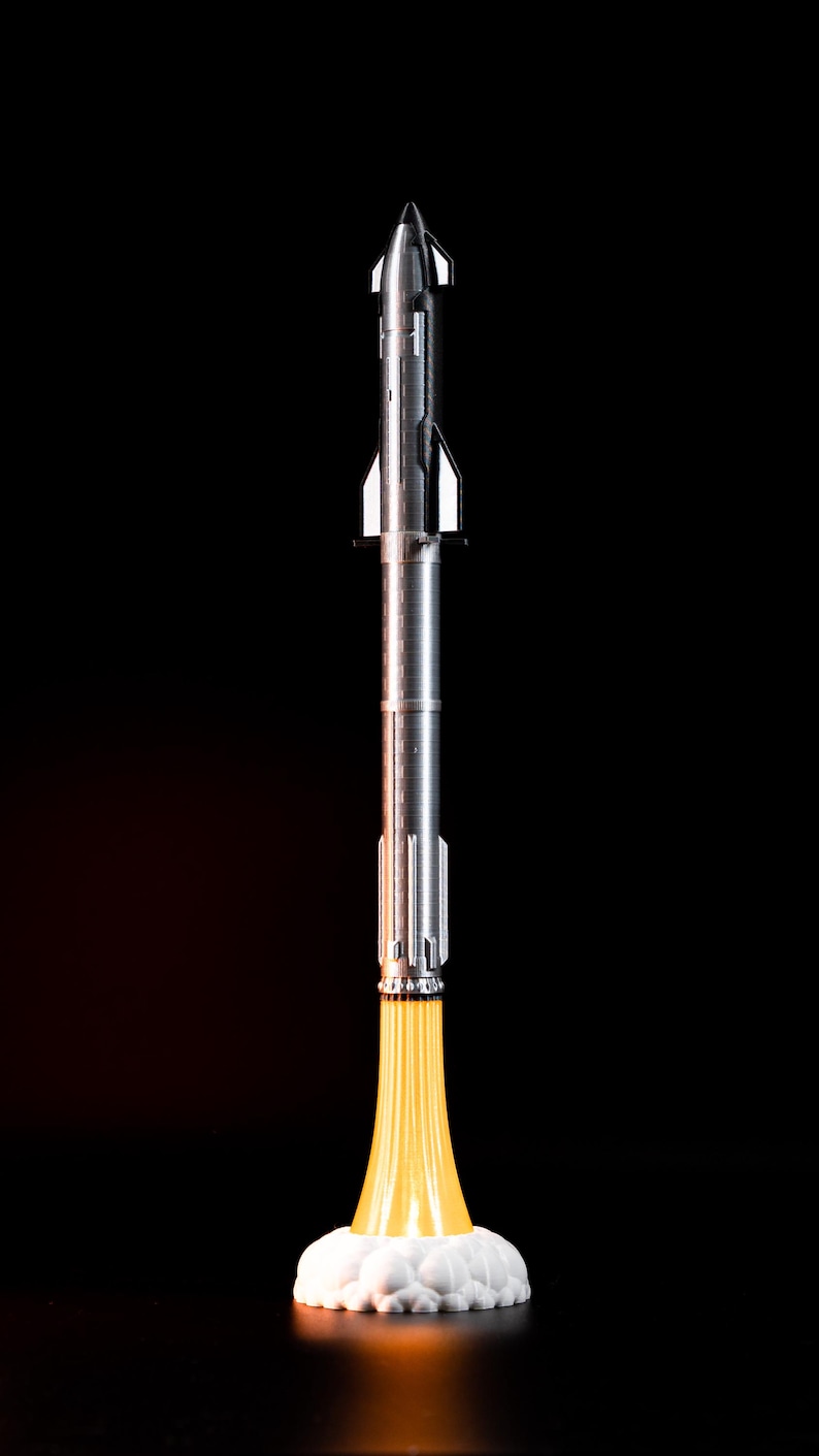 May include: A silver rocket model with a pointed nose and detailed body, emitting a bright yellow flame from its base. The rocket sits on a white cloud-like structure against a black background, creating a dramatic contrast.