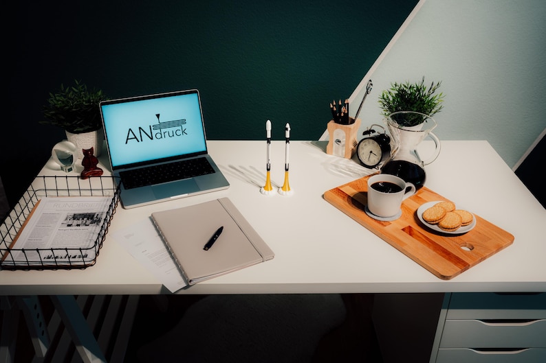 May include: A well-lit desk setup featuring a laptop with the text "ANdruck" on the screen, a notebook, pens, and a coffee break with cookies. Two rocket-shaped objects stand upright. A wire basket holds papers.