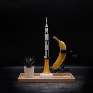 May include: A model rocket ship with black and white stripes, launching with a yellow flame, sits on a wooden tray. A banana is held in a clamp, and a small potted plant is also on the tray, all against a black background.