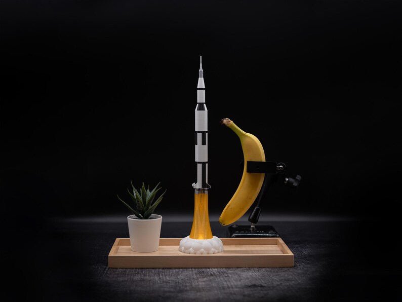 May include: A model rocket ship with black and white stripes, launching with a yellow flame, sits on a wooden tray. A banana is held in a clamp, and a small potted plant is also on the tray, all against a black background.