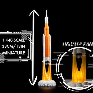 May include: A miniature rocket model, 33cm tall, with orange and white detailing. The model features an LED illuminated exhaust plume and is labeled as 1:440 scale. The miniature sits on a white cloud-like base against a black background.