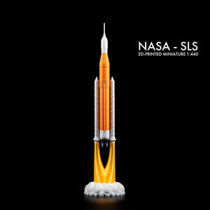May include: A 3D-printed miniature of the NASA SLS rocket, scale 1:440. The model is orange and white, with a detailed design. The rocket is depicted launching, with flames and smoke from the base. The text "NASA - SLS" is displayed.