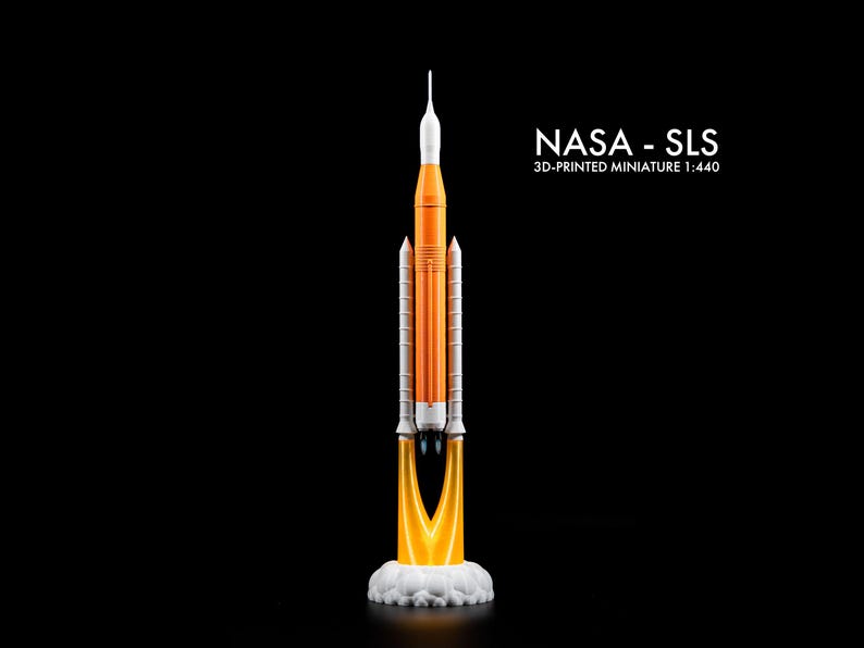 May include: A 3D-printed miniature of the NASA SLS rocket, scale 1:440. The model is orange and white, with a detailed design. The rocket is depicted launching, with flames and smoke from the base. The text "NASA - SLS" is displayed.