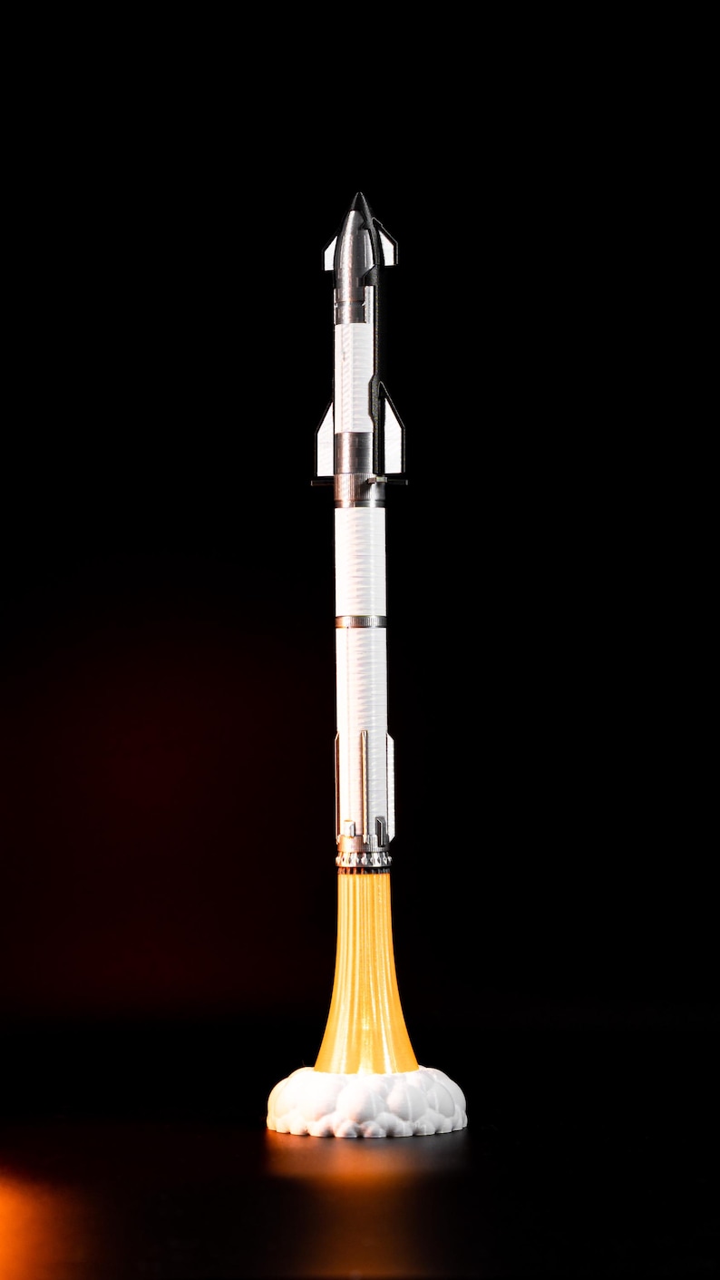 May include: A detailed model rocket with a silver and white body, set against a black background. The rocket has a pointed nose and fins, with a bright yellow flame effect emanating from the base, creating a dynamic visual.