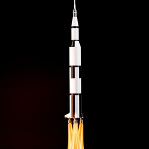 May include: A model rocket ship with a white and black body, a pointed top, and a fiery orange exhaust. The rocket is set against a black background, giving the impression of a launch.