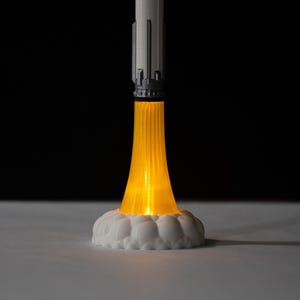 May include: A 3D-printed rocket model with a bright orange light emanating from the base, simulating flames. The rocket is white and gray, with a detailed design. The base is white and cloud-shaped, set against a black background.