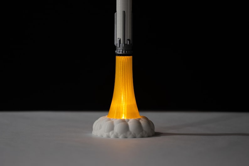 May include: A 3D-printed rocket model with a bright orange light emanating from the base, simulating flames. The rocket is white and gray, with a detailed design. The base is white and cloud-shaped, set against a black background.