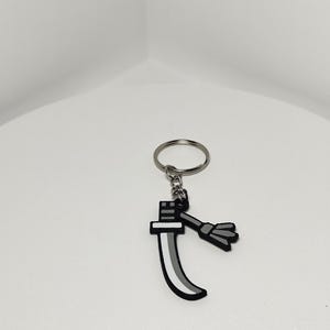 May include: A keychain featuring a stylized sword design. The sword has a black handle and a curved blade with a gray and white gradient. A small, gray tassel hangs from the handle. The keychain ring is silver.