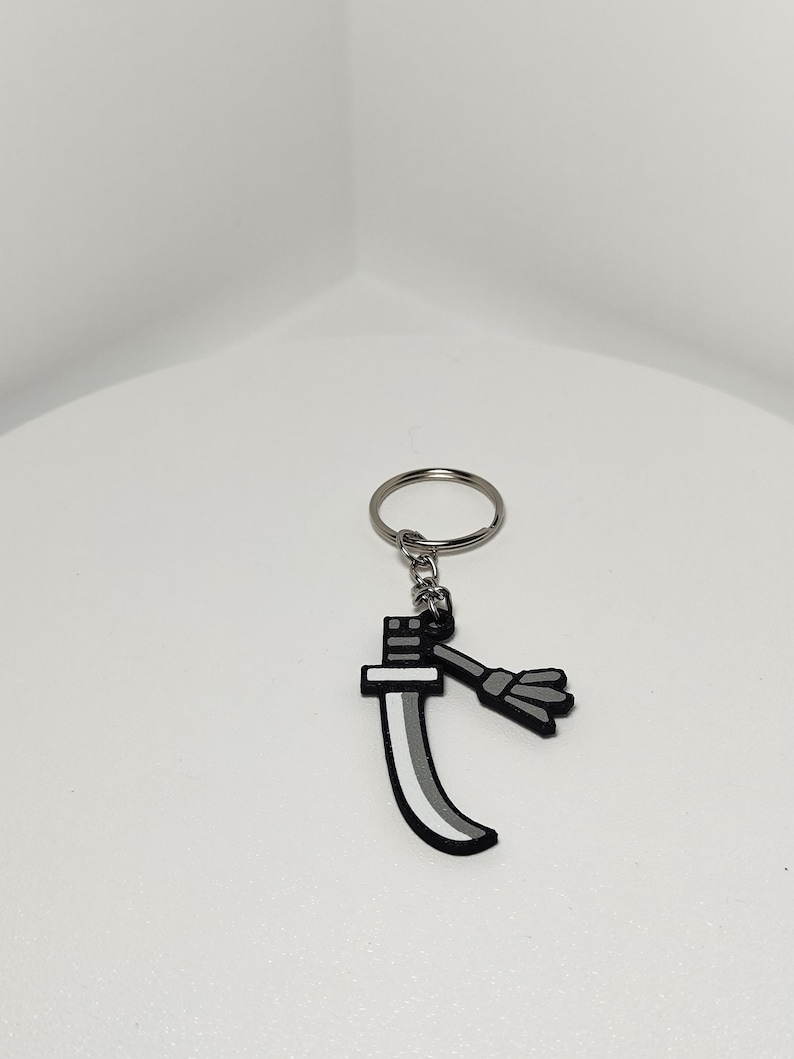 May include: A keychain featuring a stylized sword design. The sword has a black handle and a curved blade with a gray and white gradient. A small, gray tassel hangs from the handle. The keychain ring is silver.