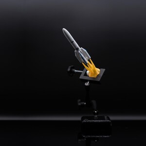 May include: A miniature rocket model, primarily white and gray, with blue accents, is depicted launching from a black base. The rocket emits a yellow flame, suggesting liftoff. The background is a solid black, emphasizing the model.
