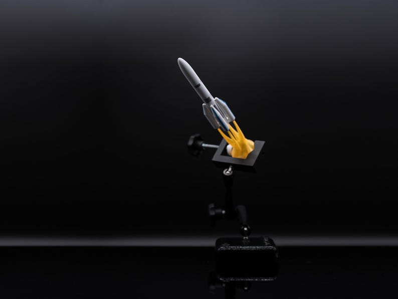 May include: A miniature rocket model, primarily white and gray, with blue accents, is depicted launching from a black base. The rocket emits a yellow flame, suggesting liftoff. The background is a solid black, emphasizing the model.