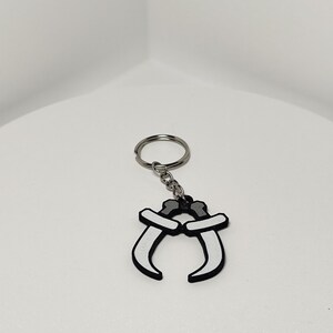 May include: A keychain featuring a black and white design of two curved blades. The blades are connected at the top with a silver-colored chain and keyring. The keychain is set against a plain white background.