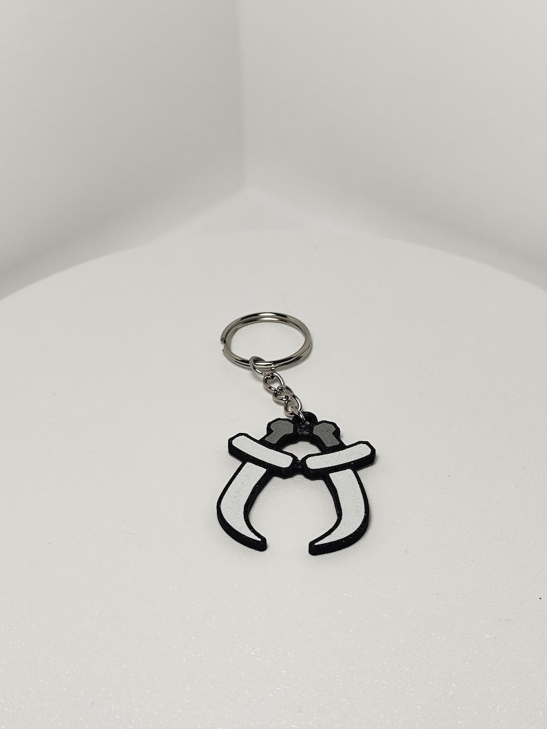 May include: A keychain featuring a black and white design of two curved blades. The blades are connected at the top with a silver-colored chain and keyring. The keychain is set against a plain white background.