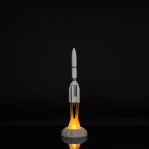 May include: A white and blue rocket model with orange flames emitting from the base, set against a black background. The rocket is standing on a gray, cloud-like base, with a bright glow illuminating the scene.