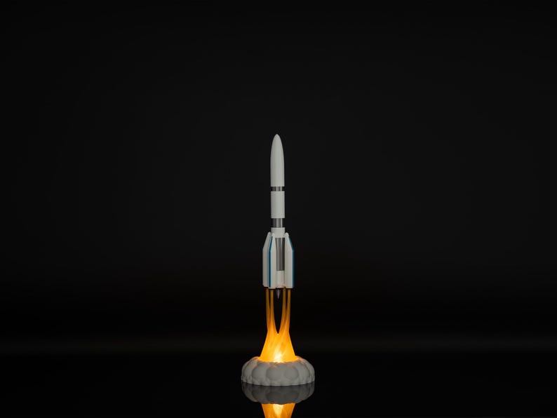 May include: A white and blue rocket model with orange flames emitting from the base, set against a black background. The rocket is standing on a gray, cloud-like base, with a bright glow illuminating the scene.