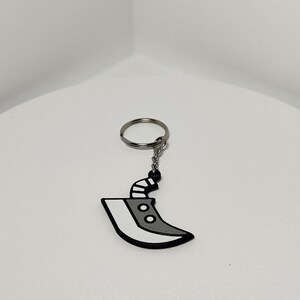 May include: A keychain featuring a stylized, gray and white blade design with a black outline. The blade has two circular details and a black handle. The keychain includes a silver-colored ring and chain.