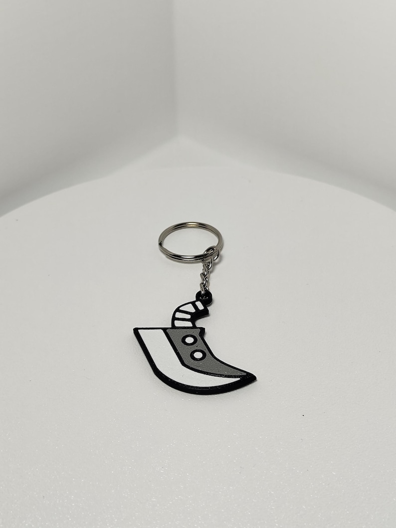 May include: A keychain featuring a stylized, gray and white blade design with a black outline. The blade has two circular details and a black handle. The keychain includes a silver-colored ring and chain.