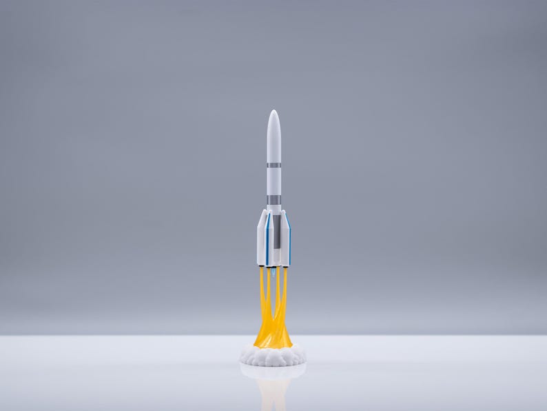 May include: A white and blue toy rocket with yellow flames blasting from the base. The rocket has a pointed top and several gray bands around the body. The rocket is on a white surface against a gray background.