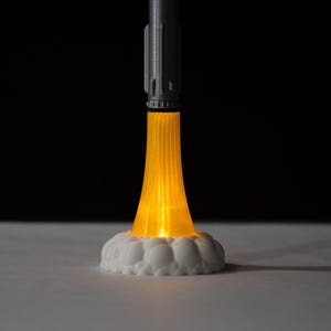 May include: A 3D-printed rocket lamp with a gray body and a bright orange light emanating from the base, resembling flames. The rocket sits on a white, cloud-like base, creating a futuristic and playful design. The background is black.