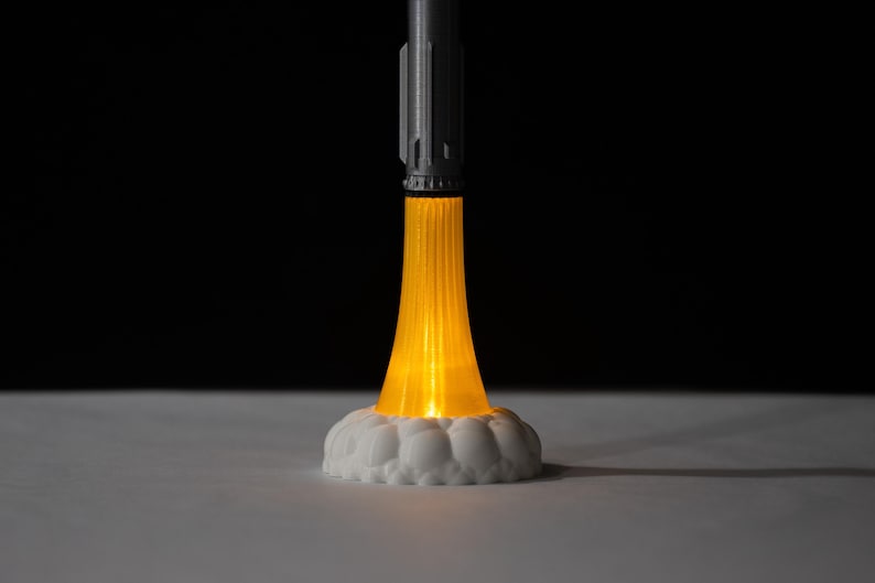 May include: A 3D-printed rocket lamp with a gray body and a bright orange light emanating from the base, resembling flames. The rocket sits on a white, cloud-like base, creating a futuristic and playful design. The background is black.