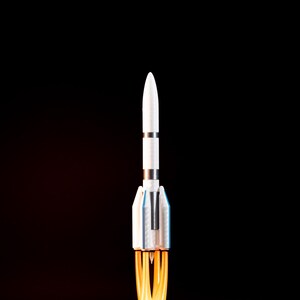 May include: A white and silver rocket model with blue accents, launching from a cloud of white smoke and orange flames against a black background. The rocket has a pointed nose and detailed body.