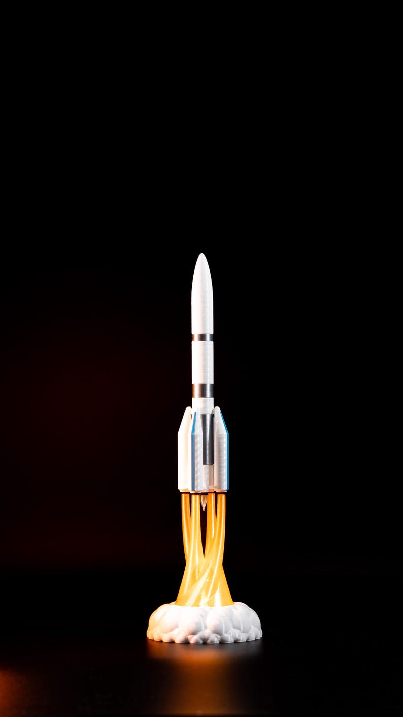 May include: A white and silver rocket model with blue accents, launching from a cloud of white smoke and orange flames against a black background. The rocket has a pointed nose and detailed body.